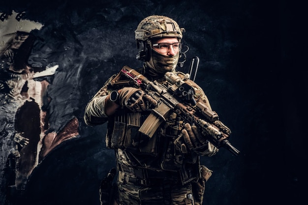 Volledig uitgeruste special forces-soldaat in camouflage-uniform met een aanvalsgeweer. Studiofoto tegen een donkere muur
