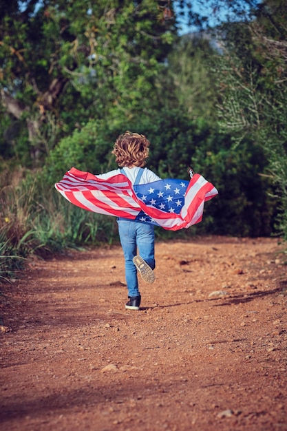 Volledige achterzijde van een anonieme actieve jongen die met een kleurrijke Amerikaanse vlag loopt op een landelijk pad op het platteland met groene bomen