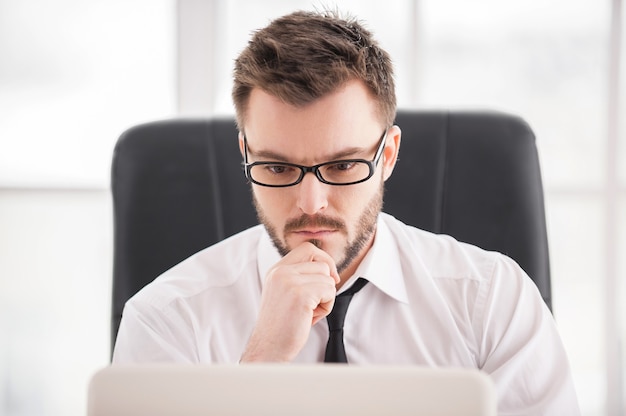 Volledige concentratie aan het werk. Knappe jonge baardman in overhemd en stropdas die op laptop werkt terwijl hij op zijn werkplek zit