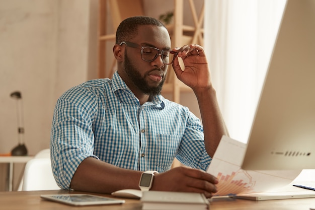 Volledige concentratie slimme jonge afro-amerikaanse man in glazen die zittend op de computer werkt