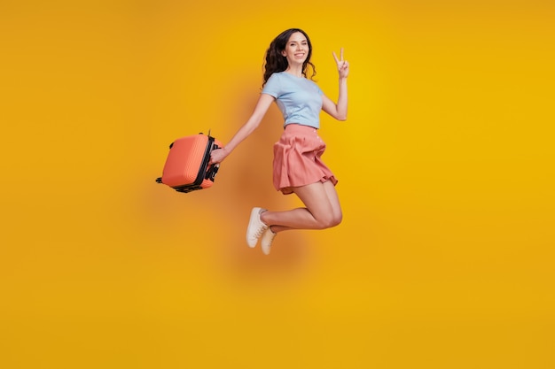 Volledige foto van jonge vrouw gelukkig positieve glimlach tas koffer springshow vrede v-teken geïsoleerd over gele kleur achtergrond