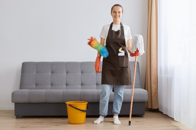 Volledige foto van lachende positieve vrouwelijke schoonmaakvloer met handschoenen, schort, t-shirt en spijkerbroek met dweilwasmiddel en ppduster voor het reinigen van stof kijkend naar de camera