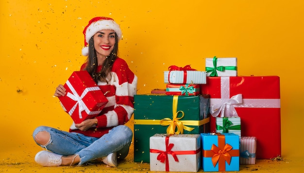 Volledige foto van opgewonden jonge lachende mooie en charmante vrouw in kerstmuts en met kerststemming terwijl ze zit met veel kleurrijke geschenkdozen en plezier heeft met confetti