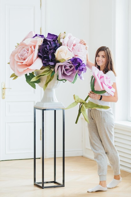 Volledige foto van tevreden brunette Europese vrouw gekleed in casual kleding maakt boeket van bloemen poses binnen in ruime kamer Jonge zakenvrouw bezit bloemenwinkel Indoor schot