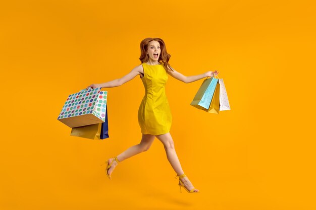 Volledige foto van verraste roodharige dame draagt veel shopping packs verslaafd shopaholic in gele jurk vreugde winkelcentrum geïsoleerd ove gele kleur achtergrond in studio