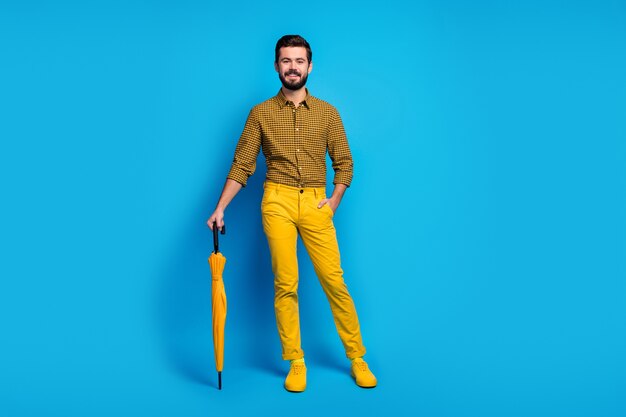 Volledige grootte foto van spontane imposante dromerige man geniet van vrije tijd rust ontspannen hebben glans paraplu draag goed uitziende moderne kleding sneakers geïsoleerd over blauwe kleur