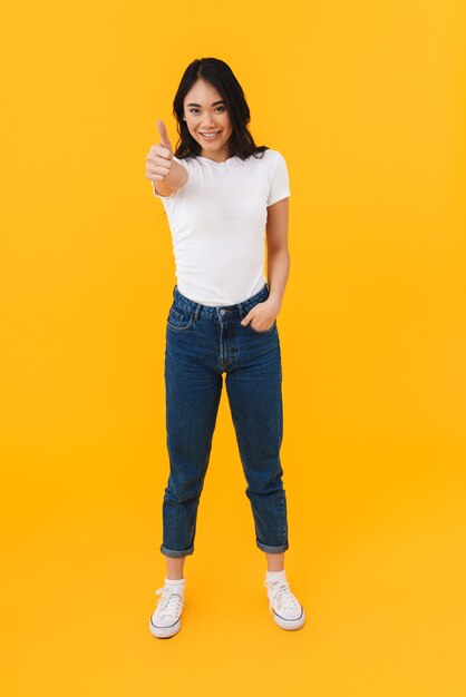 Volledige lengte afbeelding van jonge brunette aziatische vrouw glimlachend en gebaren duim omhoog geïsoleerd op geel