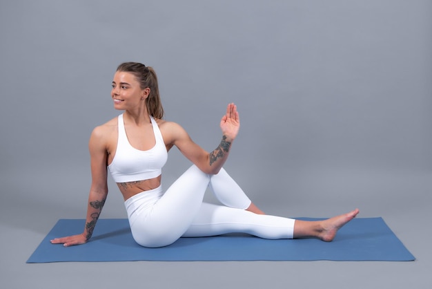 Volledige lengte fitness vrouw in witte sportkleding zittend op gym mat en het beoefenen van yoga geïsoleerd op een grijze achtergrond