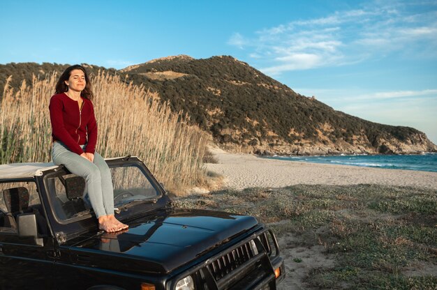 Volledige lengte Jonge vrouw zit tegen de wind op het dak van haar off-road auto met gesloten ogen. Auto geparkeerd voor een wild strand