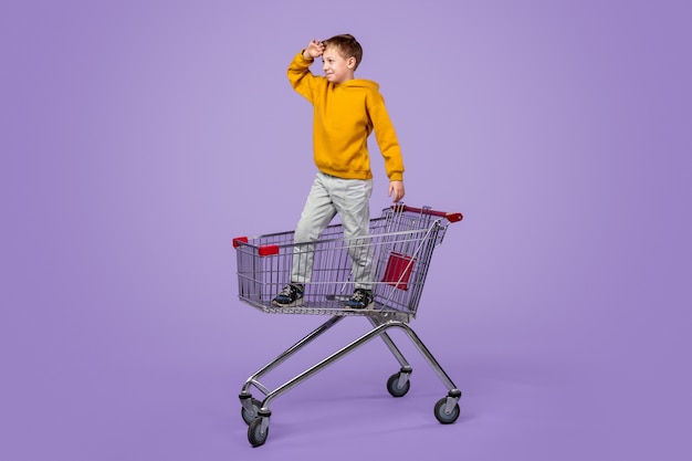 Volledige lengte jongen in casual outfit wegkijken en rijden op een trolley terwijl hij plezier heeft tijdens het winkelen tegen een paarse achtergrond