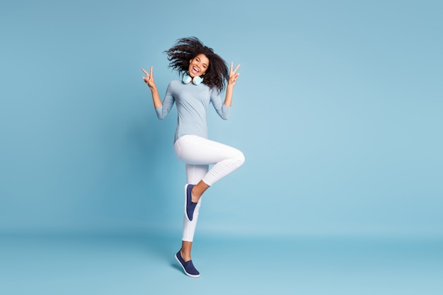 Volledige lengte lichaamsgrootte foto van vrolijke positieve mooie mooie jongere springen dansen tonen u v-sign in schoeisel witte broek geïsoleerde pastel blauwe kleur achtergrond