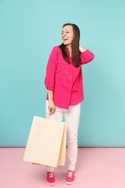 Volledige lengte portret lachende jonge vrouw in roze shirt blouse, witte broek houden boodschappentassen geïsoleerd op fel roze blauwe pastel muur.