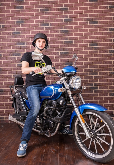 Volledige lengte portret van jonge man met helm zittend op klassieke blauwe motorfiets voor bakstenen muur