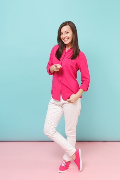 Volledige lengte portret van lachende jonge mooie vrouw in roze shirt blouse, witte broek poseren geïsoleerd op fel roze blauwe pastel muur.