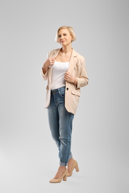 Volledige lengte portret van senior vrouw in witte tank top beige jas en vriendje jeans over grijze achtergrond