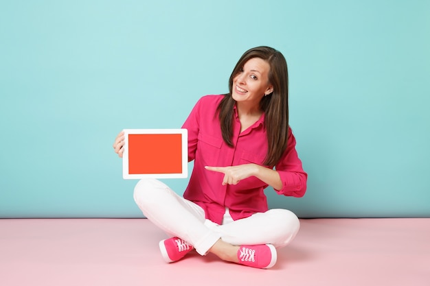 Volledige lengte portret vrouw in roze shirt blouse, witte broek zittend op de vloer houden tablet pc geïsoleerd op fel roze blauwe pastel muur.