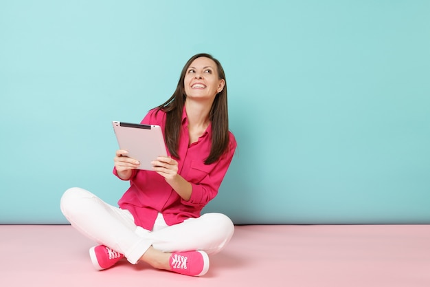 Volledige lengte portret vrouw in roze shirt blouse, witte broek zittend op de vloer houden tablet pc geïsoleerd op fel roze blauwe pastel muur.