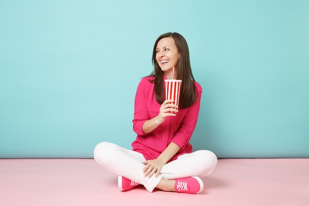 Volledige lengte portret vrouw in roze shirt blouse witte broek zittend op de vloer houdt kopje frisdrank