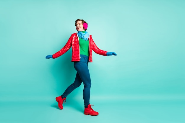 Volledige lengte profielfoto van grappige dame geniet van de winterdag actieve tijd doorbrengen lopen straatkleding casual rode overjas blauwe sjaal roze oorwarmers broek geïsoleerd groenblauw kleur muur