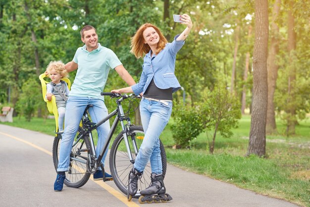 Volledige lengte shot van een jonge vrouw met skeelers poseren met haar man en baby op de fiets die een selfie maakt met behulp van slimme telefoon in de park copyspace.