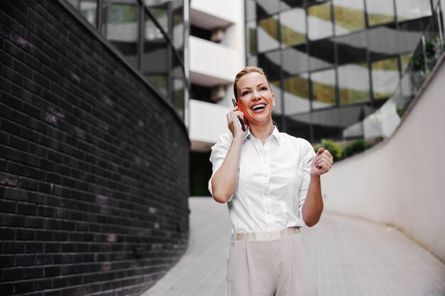 Volledige lengte van aantrekkelijke blonde modieuze onderneemster die aan de telefoon in de buitenkant van het zakencentrum spreekt.