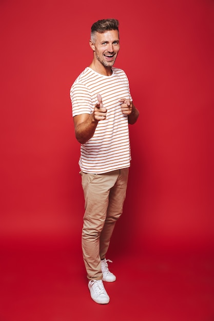 Volledige lengte van aantrekkelijke man in gestreept t-shirt gebaren wijsvingers naar je geïsoleerd op rood