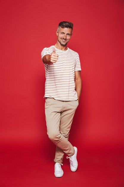 Volledige lengte van aantrekkelijke man in gestreept t-shirt glimlachend en duim opdagen geïsoleerd op rood