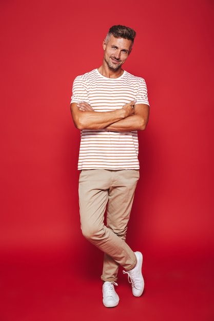 Volledige lengte van aantrekkelijke man in gestreept t-shirt glimlachend en staande met gekruiste armen geïsoleerd op rood