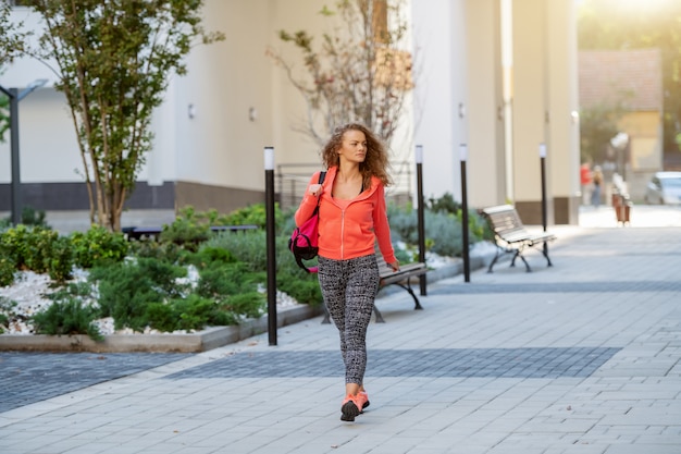 Volledige lengte van blanke vrouw in sportkleding en met krullend haar draagtas en naar de sportschool via park. Stadsleven.