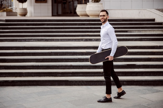 Volledige lengte van de mens in overhemd die buiten loopt en skateboard onder oksel houdt.