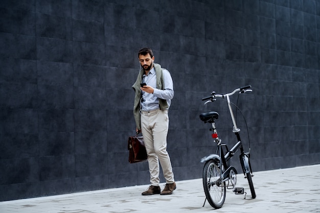 Volledige lengte van de modieuze kaukasische gebaarde aktentas van de zakenmanholding en slimme telefoon terwijl het gaan werken. in de oren zitten oortelefoons. naast de mens staat de fiets.