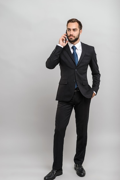 Volledige lengte van een aantrekkelijke jonge zakenman die een pak draagt dat geïsoleerd over een grijze muur staat en op een mobiele telefoon praat