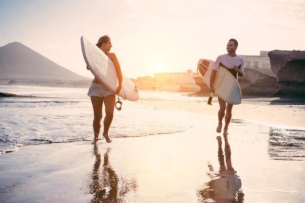 Foto volledige lengte van een jong echtpaar met een surfplank dat op het strand loopt