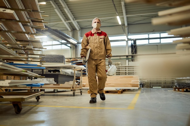 Volledige lengte van een timmerman die door een werkplaats loopt en een gezichtsmasker draagt vanwege de pandemie van het coronavirus