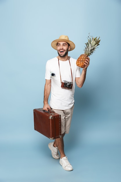 Volledige lengte van een vrolijke opgewonden man met een leeg t-shirt die geïsoleerd over de blauwe muur loopt, ananas vasthoudt, koffer draagt