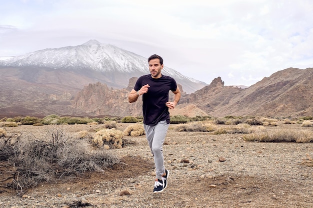 Volledige lengte van gefocuste bebaarde Spaanse mannelijke atleet in activewear die langs droog terrein in de buurt van de berg Teide rent en wegkijkt in Tenerife, Canarische eilanden, Spanje