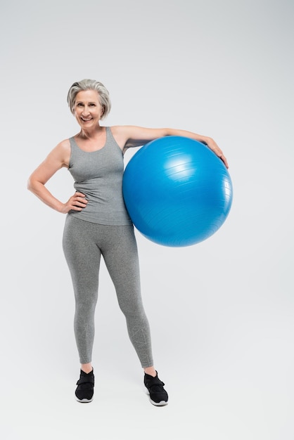 volledige lengte van gelukkige senior vrouw in sportkleding met een fitnessbal op grijs