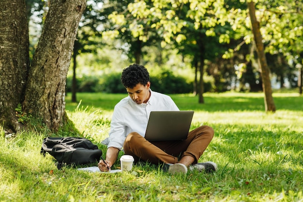 Volledige lengte van jonge man die laptop gebruikt terwijl hij in het park zit