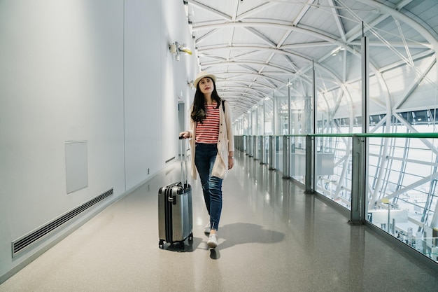 volledige lengte van jonge meisjestoerist met bagage en koffer die nieuwsgierig rondkijkt op de tweede verdieping voor vertrektijd. Gelukkige aziatische vrouw die op de luchthaven reist en loopt. reis levensstijl