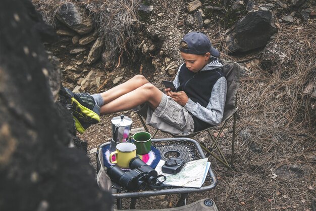 Foto volledige lengte van jongen die telefoon gebruikt terwijl hij ontspant op een stoel in het bos