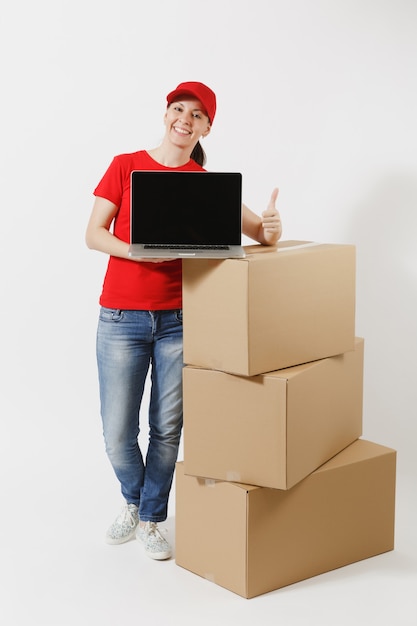 Volledige lengte van levering vrouw in rode dop, t-shirt geïsoleerd op een witte achtergrond. Vrouwelijke koerier in de buurt van lege kartonnen dozen, laptop pc-computer, leeg zwart leeg scherm. Pakket ontvangen. Ruimte kopiëren.