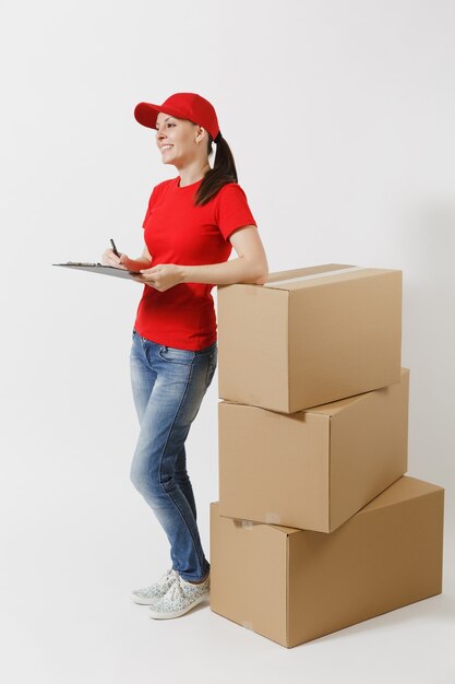 Volledige lengte van levering vrouw in rode dop, t-shirt geïsoleerd op een witte achtergrond. Vrouwelijke koerier met klembord met papieren document, leeg leeg vel op lege kartonnen dozen. Pakket ontvangen.