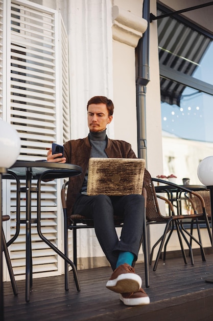 Volledige lengte van mannelijke freelancer die op mobiele telefoon surft terwijl hij op een laptop werkt en in café zit