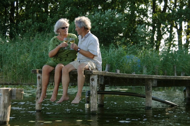 Volledige lengte van senior paar drinken terwijl ze zitten op de steiger over het meer tegen bomen