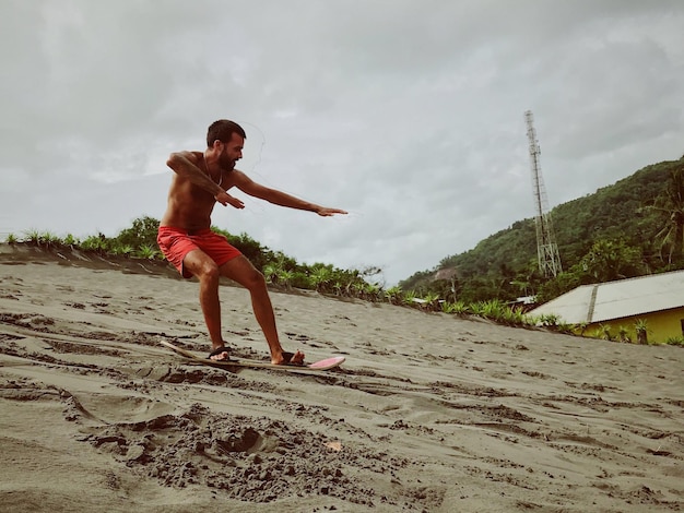Foto volledige lengte van shirtloze man die op een surfplank staat op het strand tegen de lucht.