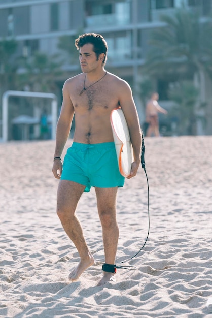 Volledige lengte van shirtloze man met surfboard die op het strand staat