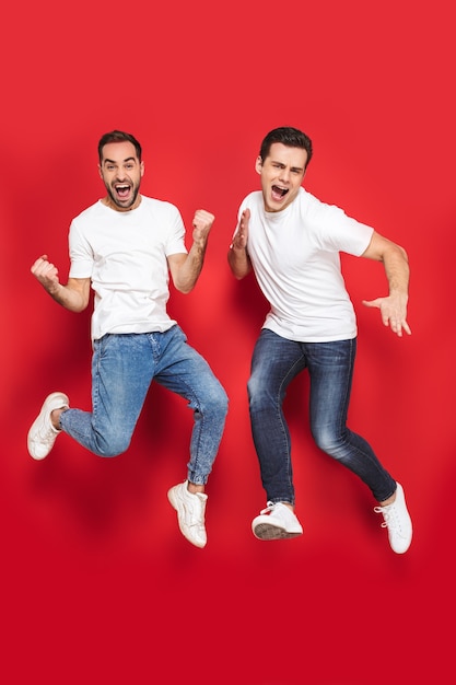Volledige lengte van twee vrolijke opgewonden mannenvrienden die lege t-shirts dragen die geïsoleerd over de rode muur springen, vieren
