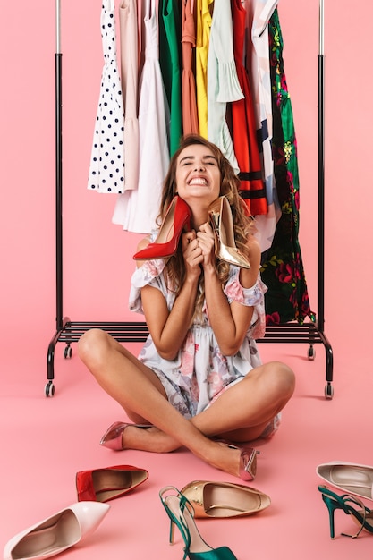 Volledige lengte van vrolijke vrouw in jurk zittend onder garderobe rek met schoenen geïsoleerd op roze