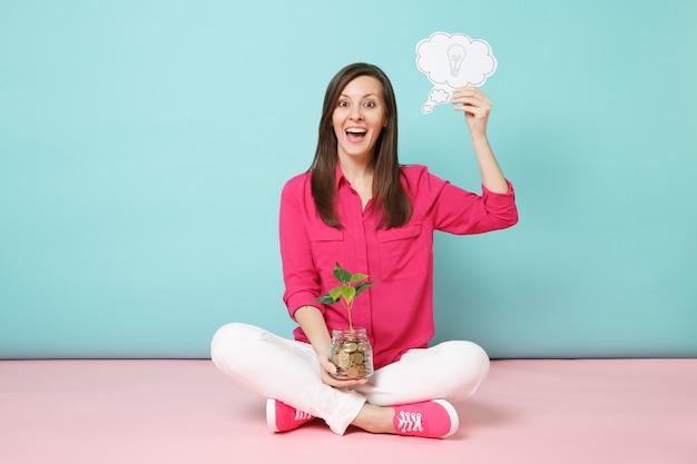 Volledige lengte van vrouw in roze shirt, witte broek zittend op de vloer met gouden munten in glazen pot