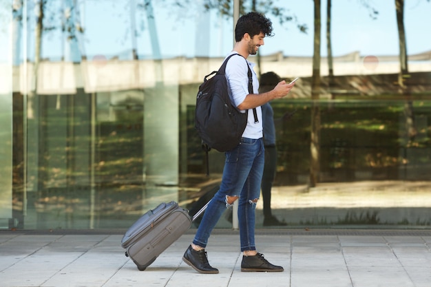 Volledige lichaams knappe mens die buiten met koffer en slimme telefoon lopen